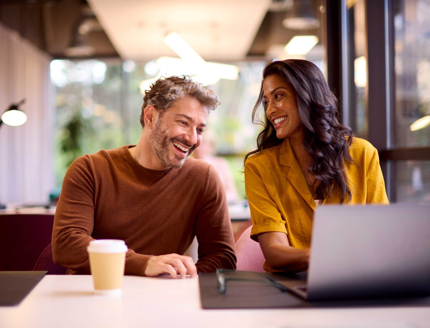 White man and asian woman in yellow shirt smilig and looking at computer while drinking coffee.